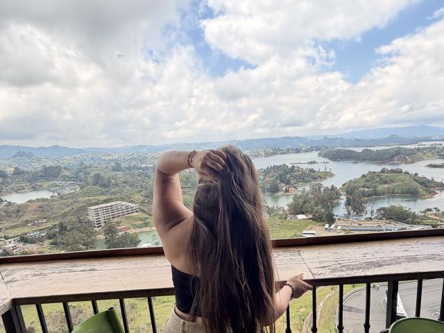 LorensDouce, back to camera, long brown hair, black top, hand in hair, standing on balcony, looking at scenic valley with lakes, green hills, and cloudy sky.