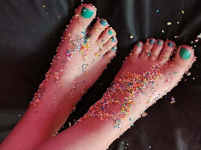 StephanieSun's feet are covered in colorful sprinkles, with teal-painted toes, on a dark surface. Sprinkles are scattered across her soles and toes.