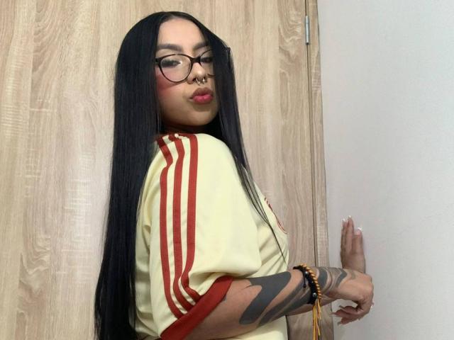 ChloeMoretti with long black hair, glasses, nose ring, and tattooed arm, wearing a yellow and red shirt, pouting, standing against a wooden door.