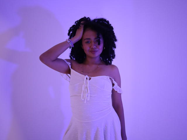 KirasPell, con cabello negro rizado, vestida con un vestido blanco de un solo hombro, con la mano en el cabello, iluminada con luz púrpura, sonriendo, con senos pequeños, con una pulsera en la muñeca, con una sombra en la pared..