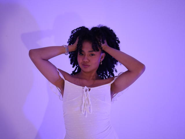 KirasPell heeft donker krullend haar, draagt een witte jurk met een open schouder, haar handen zitten in haar haar, de achtergrond is paars verlicht, ze heeft een armband om haar rechterpols, haar uitdrukking is serieus, met een lichte frons..
