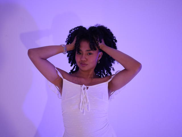 KirasPell with dark curly hair, white off-shoulder top, hands in hair, purple light, bracelet on left wrist, subtle smile, soft shadows, slight blush on cheeks.