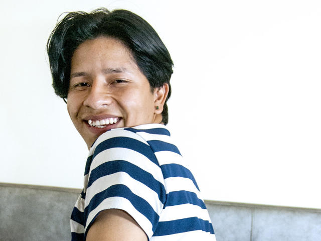 VictumMontenegro, com cabelo preto curto, sorrindo, vestindo uma camisa listrada azul e branca, olhando por cima do ombro, mostrando dentes brancos, pequenos brincos de ouro e um leve bronzeado..