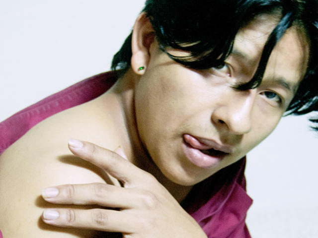 VictumMontenegro tiene el cabello oscuro, está sacando la lengua y está tocando su hombro desnudo con una mano. Lleva una camisa roja. Se ve un pendiente..