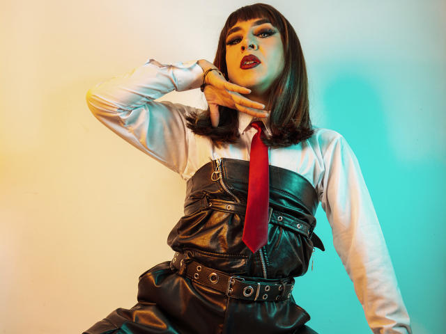 DarianaGio met een donker kort kapsel, draagt een witte blouse, een rode stropdas, een zwarte leren jurk met een riem en studs, haar hand is in de buurt van haar nek, ze kijkt omhoog met een zelfverzekerde, sexy uitdrukking..