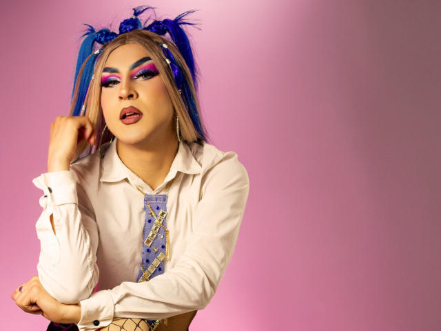 DarianaGio met blauwe vlechtjes, een wit shirt en kleurrijke make-up, met haar tong uitgestoken, een hand in de buurt van haar gezicht, een geruit gestreept das aan, er sexy uitzien, een roze achtergrond met een kleurovergang..