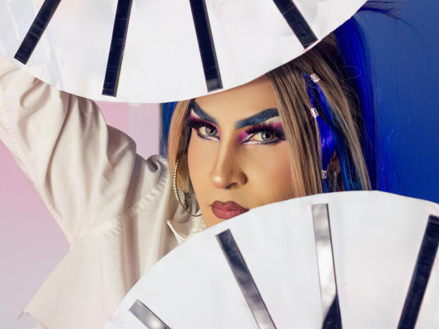 DarianaGio met opvallende blauwe oogschaduw, roze eyeliner en blauw haar, die witte waaier met zwarte strepen vasthoudt, een witte blouse draagt en gouden oorbellen heeft, en een intense blik heeft..