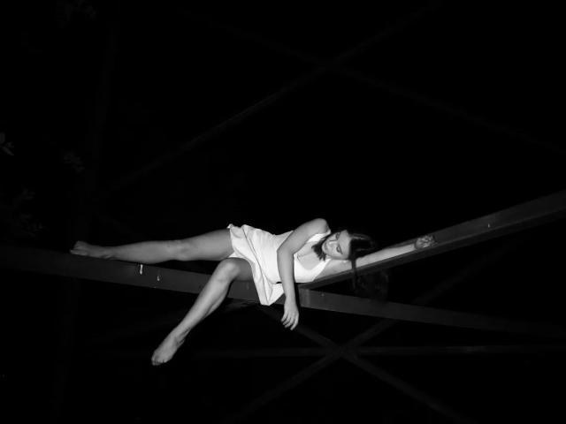 RosendaLazcano is lying on a metal beam, wearing a white dress, with her legs stretched out and arms crossed, in a dark, black-and-white photo. Her ass is prominent.