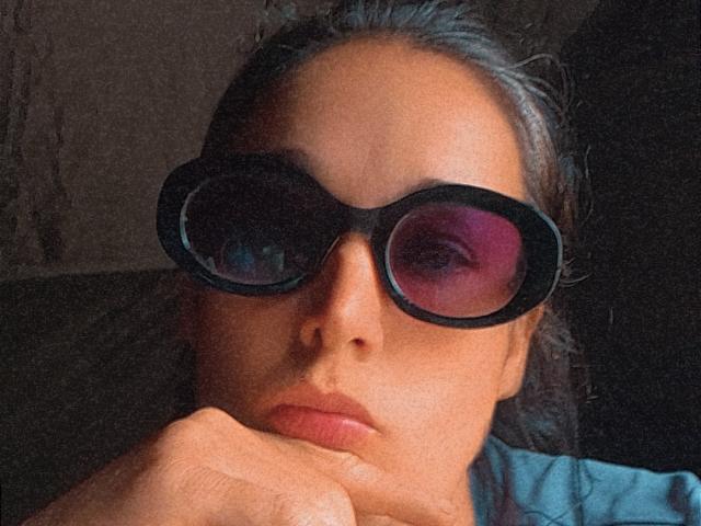 AdelineGill with huge black 3D glasses, pink reflection in lenses, dark hair, pouty lips, resting chin on hand, serious expression, light brown skin, subtle makeup.