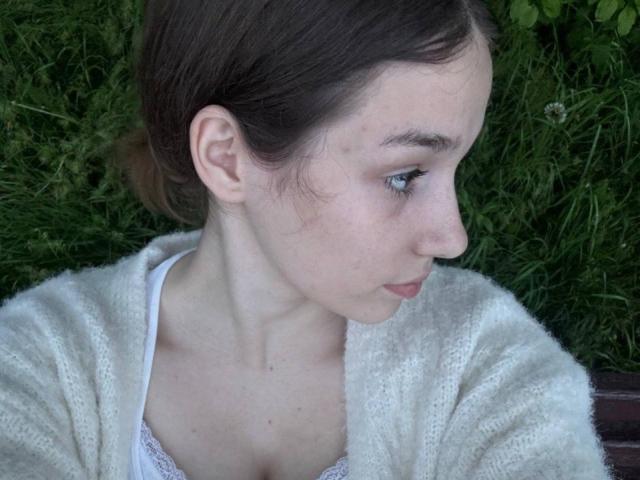 KaiaLein has pale skin, brown hair in a ponytail, and is wearing a white fuzzy cardigan with a white lace top. She's looking down with blue eyes, slightly pouty.