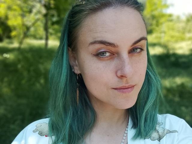 RayeReam, con i capelli verde acqua, lentiggini e occhi verdi, guarda di lato con un sorriso appena accennato, indossa una maglietta bianca con disegni di pecore e una collana di perle..