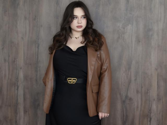 MaddyCroft, avec ses cheveux bruns ondulés, porte une veste en cuir marron par-dessus une robe noire avec une ceinture dorée, et se tient devant un mur en bois. Elle a une expression sérieuse..