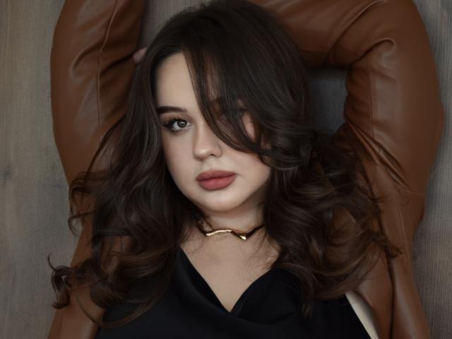 MaddyCroft, com cabelo castanho ondulado, vestindo uma blusa preta e jaqueta de couro marrom, braços acima da cabeça, olhando para a câmera com uma expressão sutil e sensual, usando um colar de ouro..