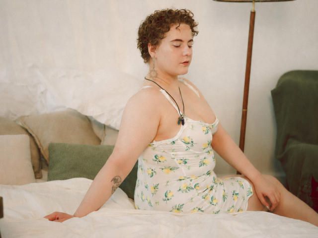 MelittaAdams with curly brown hair, white floral dress, small tits, tattoo on left arm, necklace, sitting on bed, arm behind, green pillow, white sheets, relaxed, soft lighting.