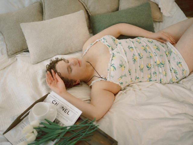 Melitta Adams liegt auf einem Bett, die Augen geschlossen, trägt ein blumiges Top, eine Hand auf dem Kopf, vor ihr auf einem Tablett ein Buch und Blumen. Sanft, entspannt, süß..