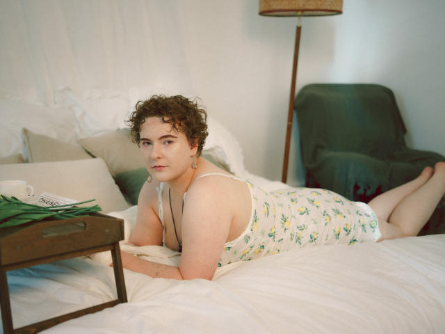 Melitta Adams, cheveux bruns bouclés, peau pâle, portant une robe blanche à motifs floraux, allongée sur un lit, les bras sous le menton, les jambes pliées, une couverture verte sur une chaise, un plateau en bois avec des feuilles vertes..