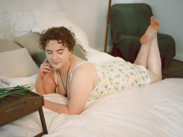 Melitta Adams, con cabello castaño rizado, piel pálida, pequeños senos, vestida con una camiseta blanca con estampado floral y pantalones cortos, acostada en una cama blanca, leyendo una revista, una silla verde al fondo, una mesa de madera con hojas verdes..