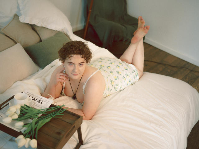 Melitta Adams, com cabelos castanhos cacheados, pele clara e seios pequenos, deitada em uma cama em um vestido branco com estampas florais, com os pés levantados, segurando uma caixa Chanel, folhas verdes, tulipas brancas..