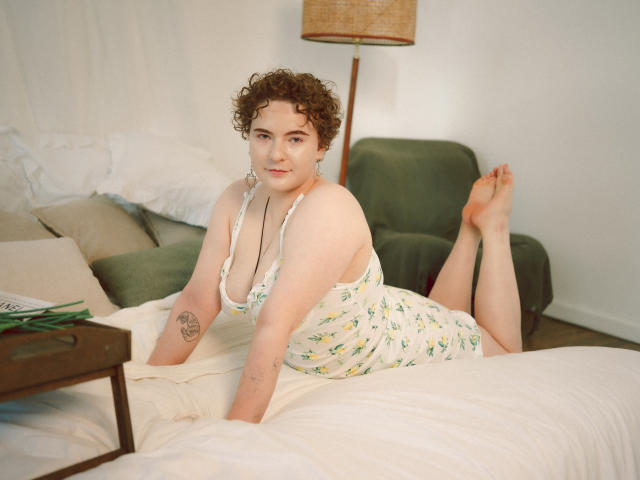 Melitta Adams, con i capelli castano ricci, la pelle chiara e piccoli tatuaggi, sdraiata su un letto con un abito bianco a fiori, che mostra il seno, con i glutei sollevati e i piedi in aria..