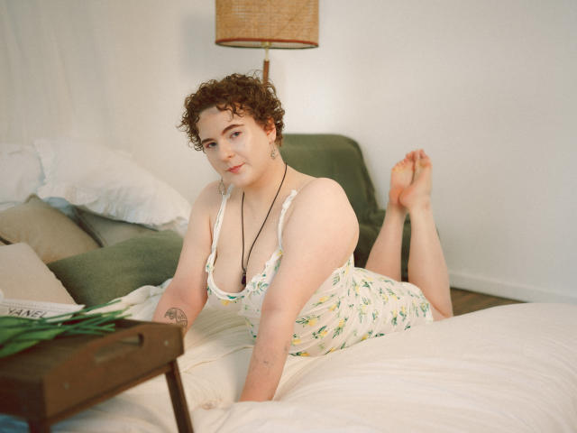 MelittaAdams with curly brown hair, white floral dress, and necklace, lying on bed, legs up, showing her ass, tattoos on arm, green pillow, wooden tray with green leaves.