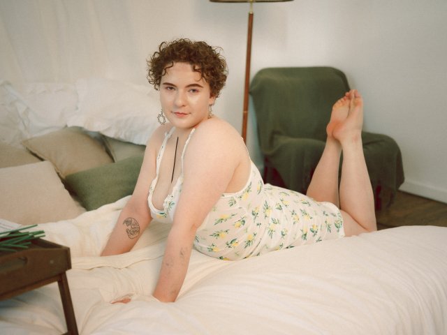 MelittaAdams with curly brown hair, wearing a floral white dress, lying on a bed, arms and legs up, showing a tattoo on her right arm, and a necklace.