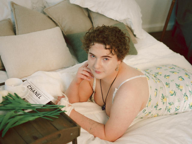 Melitta Adams ha i capelli castano ricci, la pelle chiara e sta sdraiata su un letto, indossa una canotta a fiori e guarda la fotocamera con un leggero sorriso, appoggiandosi alla mano..
