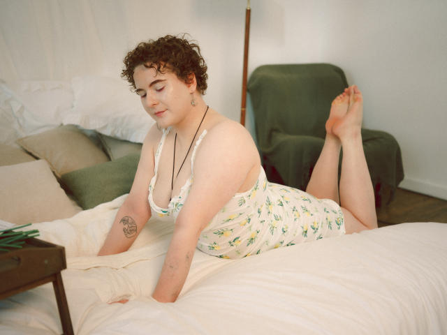 Melitta Adams, con i capelli castano ricci, la pelle chiara e piccoli seni, sdraiata su un letto con un abito bianco a fiori, mostra un tatuaggio sul braccio sinistro, con i piedi sollevati..