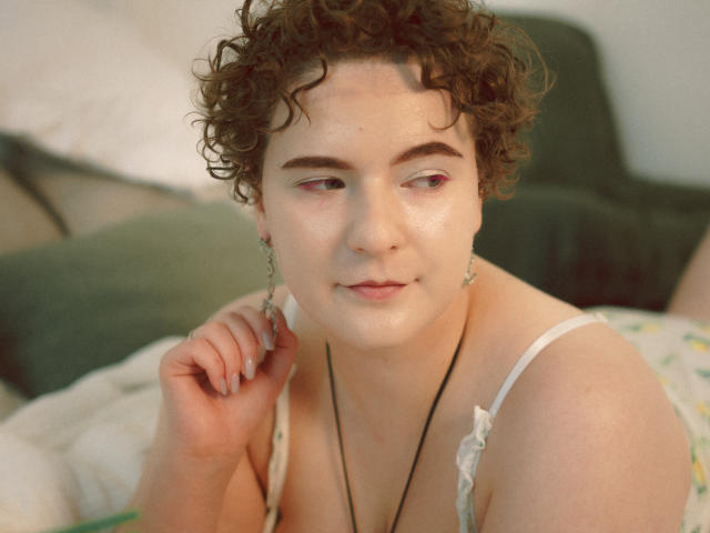 Melitta Adams, com cabelos castanhos cacheados, pele clara e pálpebras rosadas, vestindo um vestido branco com alças, usa um colar preto e está sorrindo levemente, com uma mão perto do rosto..