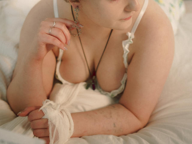 MelittaAdams with pale skin, white lace lingerie, showing big boobs, holding a white cloth, ring on finger, necklace, dangle earrings, lying on bed, looking down, tattoo on arm.