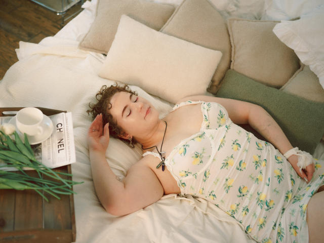 Melitta Adams, capelli castani ricci, vestito bianco con fiori, dorme su cuscini beige, letto bianco, braccia alzate, occhi chiusi, collana, libro, tazza bianca, foglie verdi, pavimento di legno, rilassata..