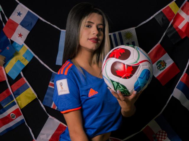 SheylaSpooner with long dark blonde hair, blue soccer shirt, holding colorful soccer ball, flags in background, smiling with red lipstick. Boobs are small, shirt has red stripes.