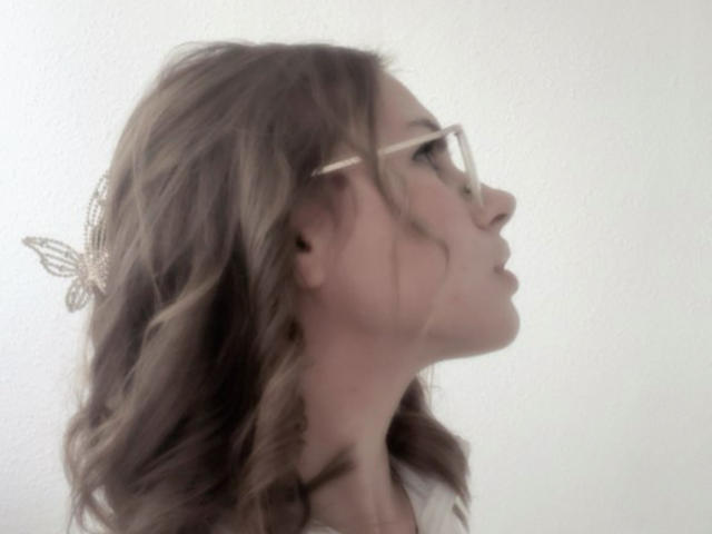HelenRosy's side profile with wavy brown hair, glasses, and a butterfly hairpin, looking up, wearing a grey top, against a plain white background.