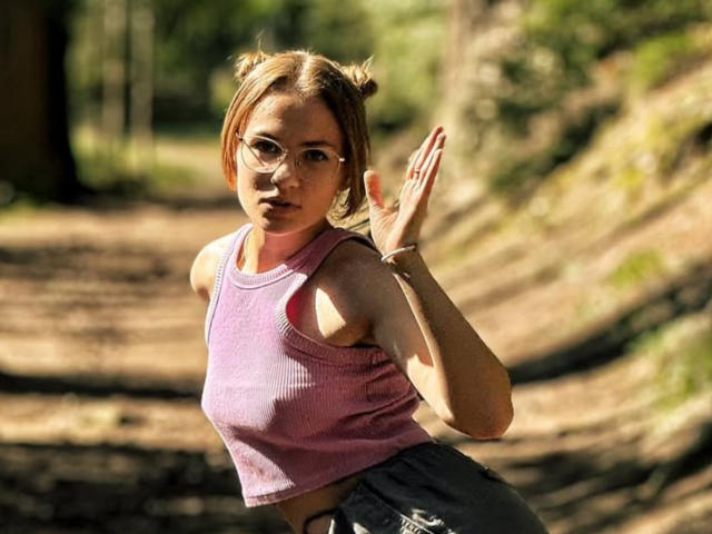 HelenRosy mit Brille, einem rosa gerippten Tanktop und grauen Shorts, mit hochgestellten Händen, überrascht in einem sonnendurchfluteten Wald. Ihre Brüste sind klein, und sie hat ein süßes, nerdiges Aussehen..