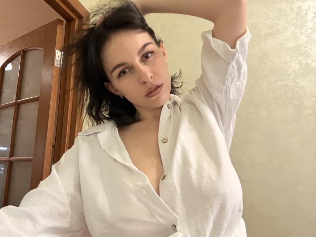 EmmaNoa, avec ses cheveux foncés, porte une chemise blanche à boutons, les bras levés, exposant ses seins, et regarde l'appareil photo avec un léger froncement de sourcils. Sa chemise est légèrement ouverte..
