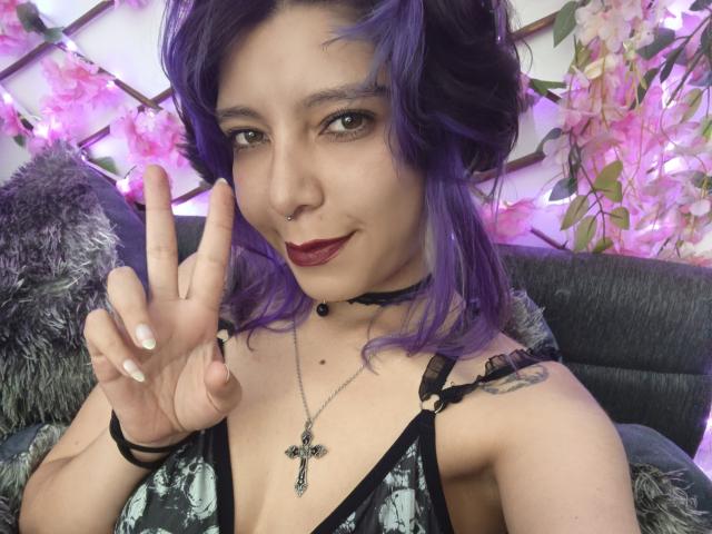 DarckVelvet with purple hair, nose ring, and black choker, showing a peace sign, wearing a black lace top with a cross necklace, smiling with dark red lipstick.