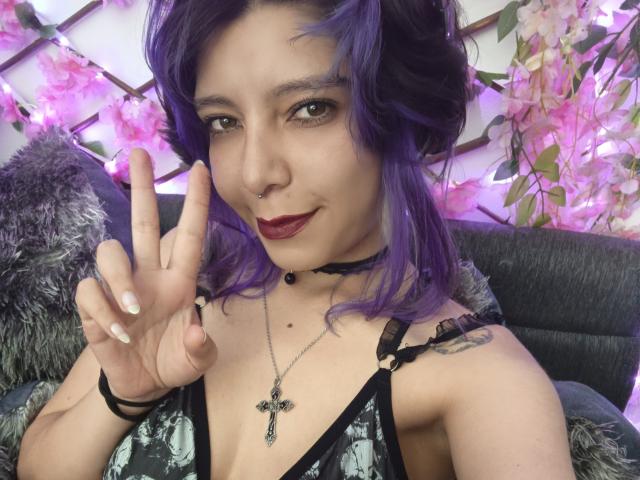 DarckVelvet mit lila Haaren, dunklem Lippenstift, Nasenring und Kreuzanhänger, die ein Friedenszeichen zeigt, trägt ein schwarzes Spitzenoberteil und hat ein Tattoo an der rechten Schulter..