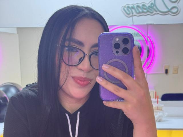 Camille, met lang zwart haar, een bril en roze lippenstift, houdt een paarse telefoonhoes vast en maakt een selfie in een lichte kamer met roze neonverlichting..