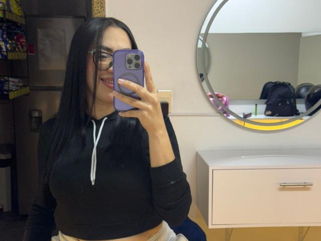 Camille, con lunghi capelli neri, occhiali, felpa nera e fascia bianca, sta scattando un selfie allo specchio con un telefono viola, mostrando il seno e sorridendo..
