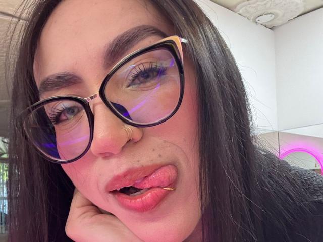 Camille, avec des lunettes noires, un anneau au nez et un piercing aux lèvres, la langue tirée, montrant ses lèvres roses, une main sous le menton, des cheveux bruns foncés et une peau claire. Ses yeux ont des reflets violets..