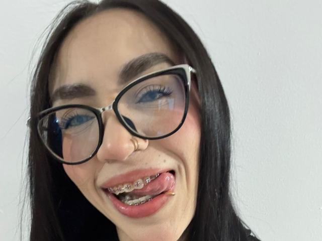 Camille, con occhiali neri, piercing al naso e lingua con griglie metalliche, sorride con occhi azzurri e capelli neri lisci, mostrando un aspetto carino ma allo stesso tempo audace..