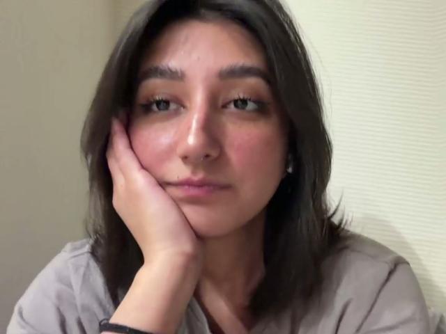 Heike Carchi, mit schulterlangem braunen Haar, grünen Augen und einem leichten Lächeln, die ihre Wange in ihrer Hand ruhend hat, trägt ein graues Hemd und hat ein dezentes Nasenpiercing..