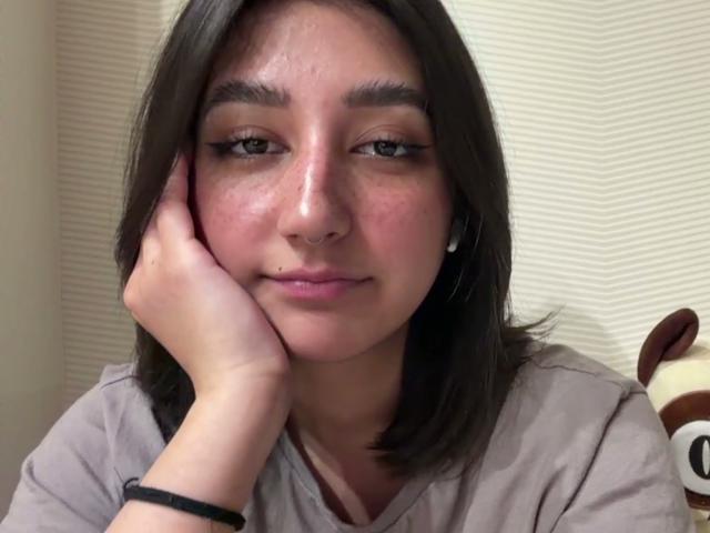 Heike Carchi tiene un rostro lindo, ligeramente sonrojado, con cabello castaño, ojos verdes y un pequeño piercing en la nariz. Descansa su mano en su mejilla, vistiendo una camisa gris..
