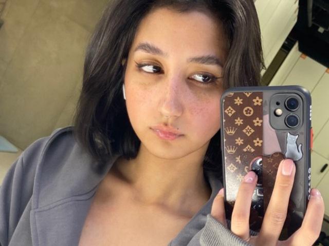 Heike Carchi, con piel de tono marrón medio, cabello oscuro y ondulado, y un piercing discreto en la nariz, sosteniendo un teléfono con una funda marrón y estampada, vistiendo una sudadera gris, mirando hacia un lado..