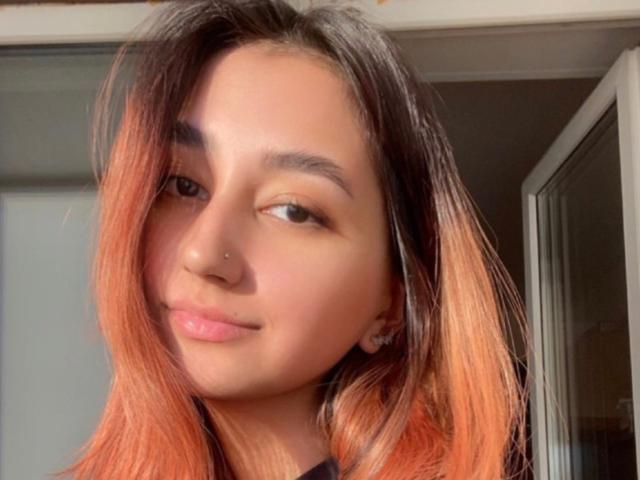 HeikeCarchi heeft een lichtbruine huid, een klein neuspiercing, een subtiele glimlach en oranje haar. Ze heeft een lichte grijns en er is zacht licht op de achtergrond. De borsten van HeikeCarchi zijn niet zichtbaar..