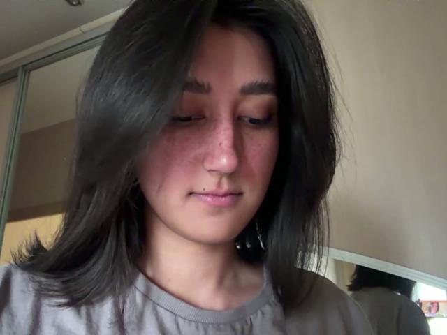 HeikeCarchi has a pimple on her nose, dark straight hair, light pink freckles, and a small lower lip piercing. She's wearing a gray shirt, looking down.