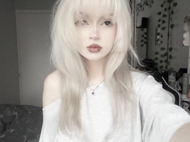 Hortencia Vergin, avec sa peau pâle, ses cheveux blonds platine, sa frange, sa chemise blanche à épaules dénudées, son rouge à lèvres foncé, son anneau au nez et son collier, prend un selfie. Ses seins sont petits..