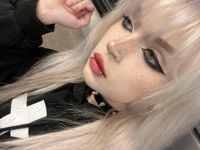 HortenciaVergin, avec une peau pâle, des taches de rousseur, des cheveux blonds platine, un eye-liner noir, du rouge à lèvres rouge, un piercing au nez, un piercing aux lèvres, un collier noir, une chemise noir et blanc, regardant la caméra, une main levée..