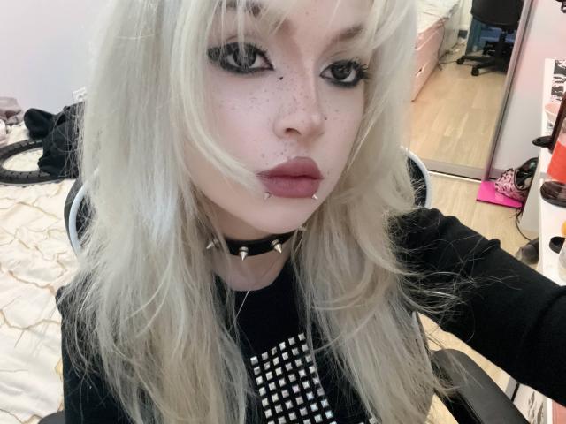 HortenciaVergin with platinum blonde hair, black eyeliner, freckles, pierced nose and lower lip, black choker, and spiked shirt, sitting in a chair, looking at the camera.