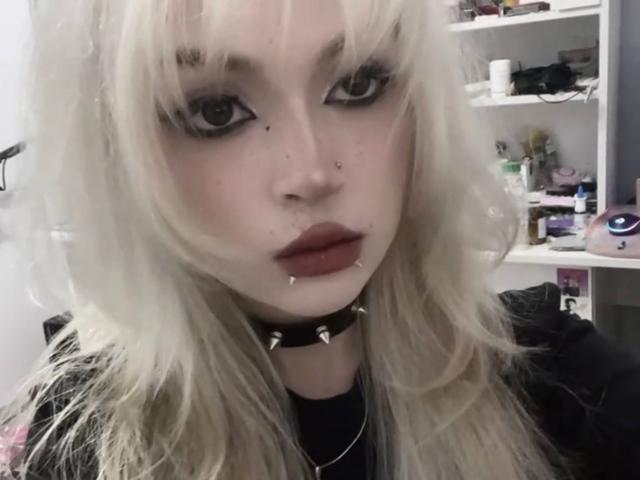 Hortencia Vergin, com cabelo loiro platinado, batom escuro, piercing no nariz e colar com espinhos, olhando para a câmera com delineador marcante e a boca ligeiramente entreaberta..