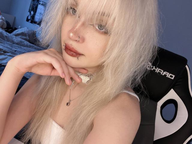 HortenciaVergin with white hair, nose ring, lip piercing, and choker, biting her finger, wearing a white top, sitting in a black and white chair. Blue-eyed, pale skin.